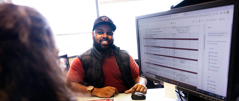 An african american male advisor sits behind a desk and is looking at a computer screen featuring an academic map with his female student.