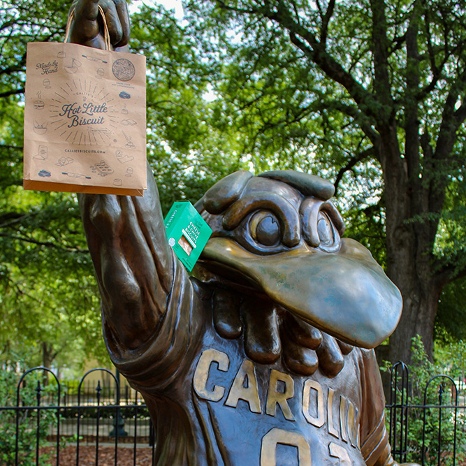 Cocky mascot statue holding a Callie’s Hot Little Biscuit bag outdoors beneath leafy trees.