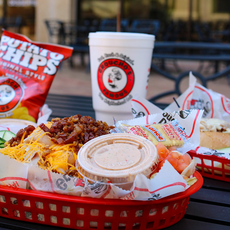 Loaded chili sandwich with bacon and cheese in a red basket, served with dipping sauce, chips, and a drink.