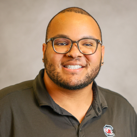 Professional headshot of a smiling person with closely cropped hair, a neatly trimmed beard, and black rectangular eyeglasses wearing a black collared polo shirt with a university logo. The person is facing the camera and has a small nose ring and stud earrings. The photo is set against a soft gray studio background with warm, even lighting, creating a friendly, approachable, and professional appearance.