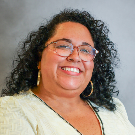 Professional headshot of a smiling person with shoulder-length curly dark hair wearing clear-framed eyeglasses, large gold hoop earrings, and a cream-colored textured short-sleeve blouse with black trim. The person is facing the camera and is wearing a small nose ring. A tattoo is partially visible on one forearm. The photo is set against a soft gray studio background with even lighting, creating a friendly, approachable, and professional appearance.