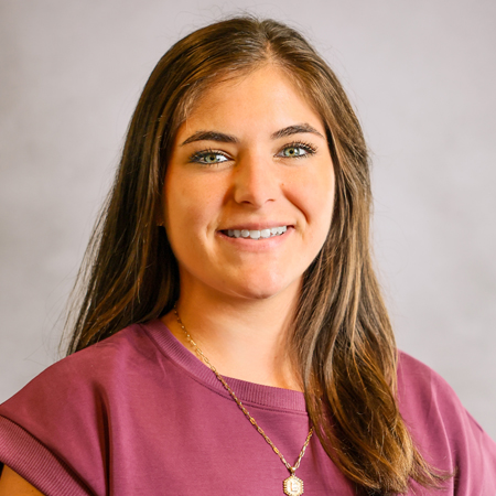 Professional headshot of a smiling person with long brown hair wearing a mauve short-sleeve blouse and a delicate gold necklace with a pendant. The person is facing the camera against a soft gray studio background with warm, even lighting, creating a friendly, approachable, and professional appearance.
