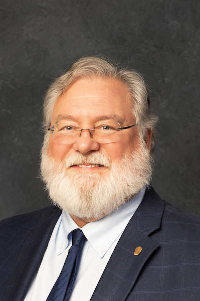 a man with a gray beard and hair wearing glasses, with a blue tie and gray jacked on standing outside