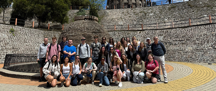 A group of University of South Carolina students and faculty pose for a photo during a semester-long study abroad trip in the Galápagos Islands, standing in front of a stone monument on a sunny day.