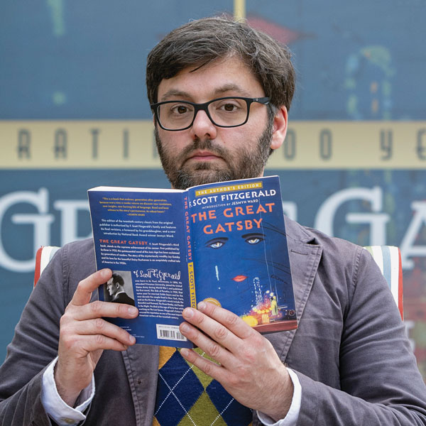Person wearing glasses reads The Great Gatsby by F. Scott Fitzgerald while looking at the camera.
