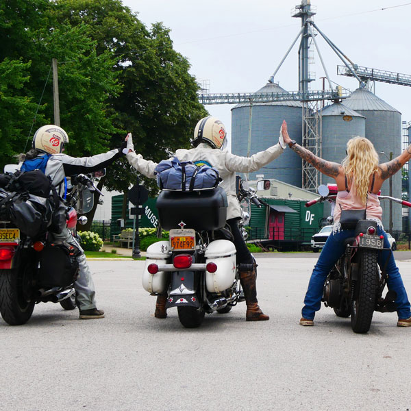 A back view of three female motorcyclists side by side, touching hands in the air
