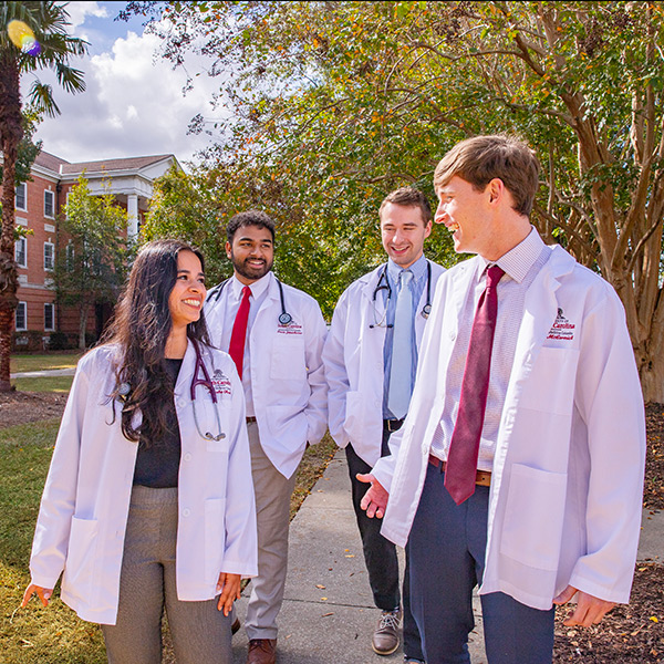Four medical students in white lab coats walk and talk on a campus pathway lined with trees and academic buildings.