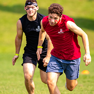 Two college-aged athletes run in competition in an open field in the heat.