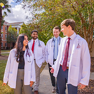 Four medical students in white lab coats walk and talk on a campus pathway lined with trees and academic buildings.